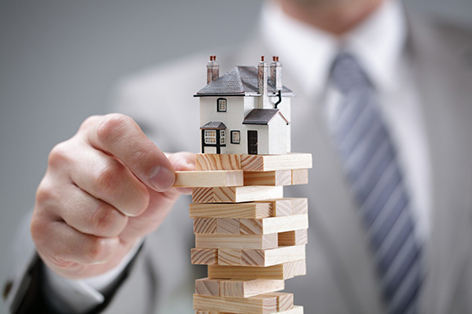 A man balancing a figurine of a house atop a precariously stacked stack of wooden blocks.