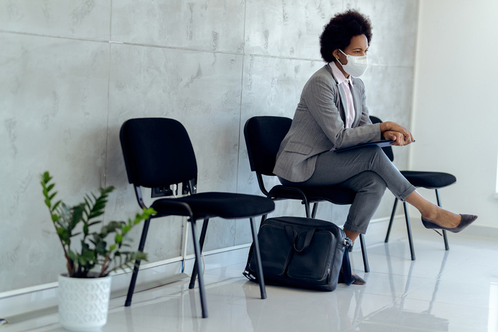 A pensive Black woman with a face mask awaiting a job interview.